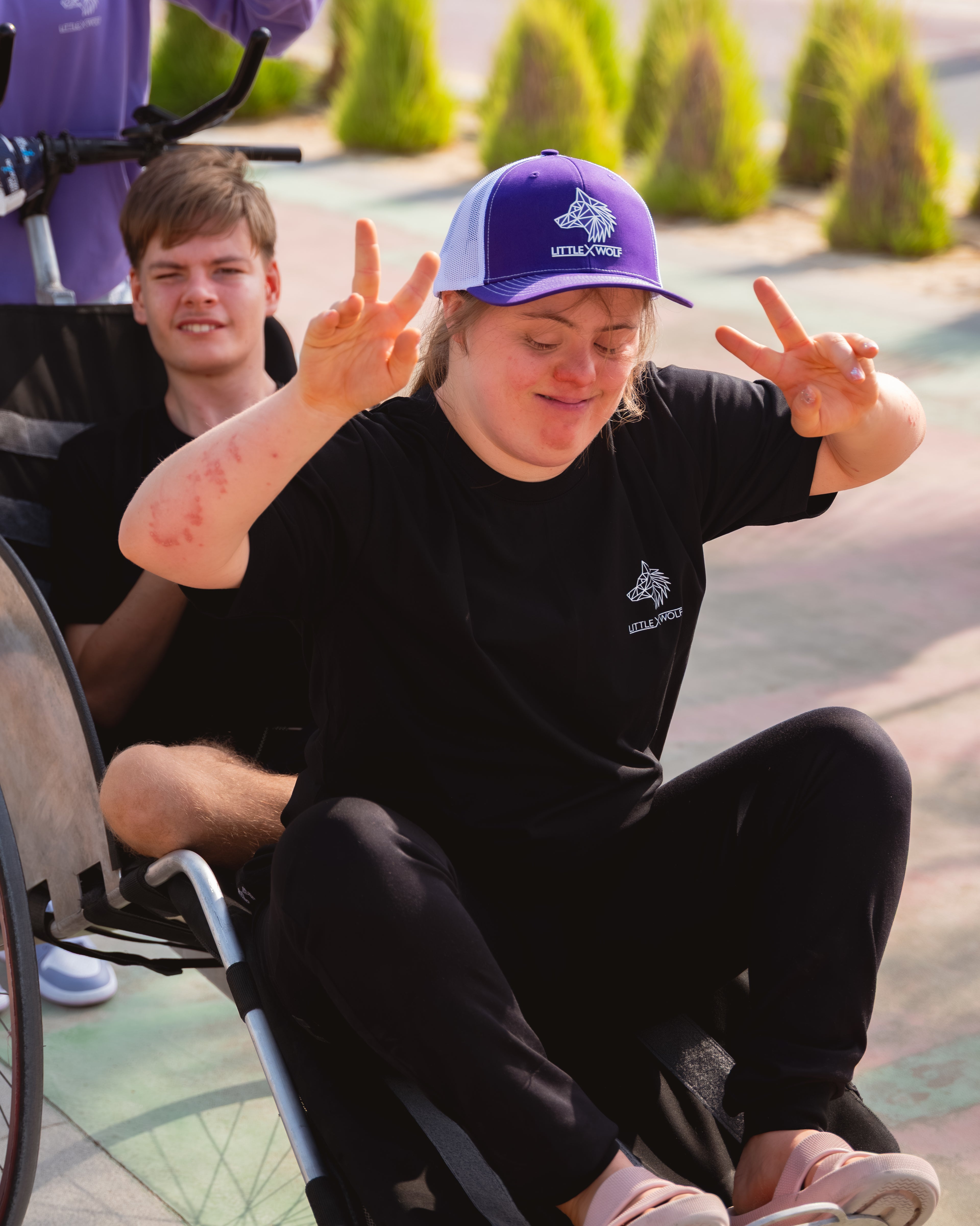 Ruby and Rio individuals in wheelchairs outdoors, one making a peace sign gesture.