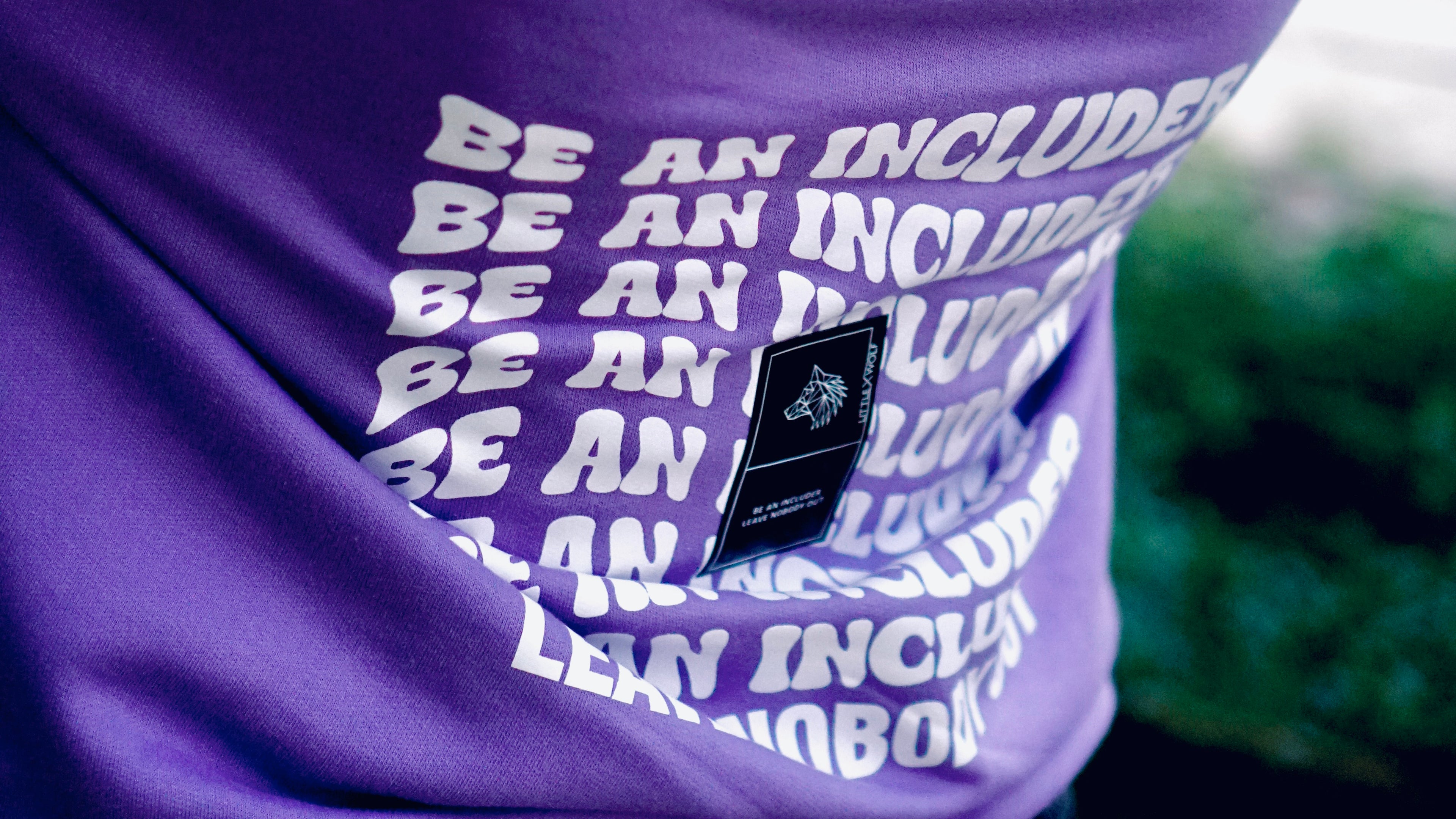 Purple hoodie with “Be An Includer” text and LittleXWolf wolf logo label.