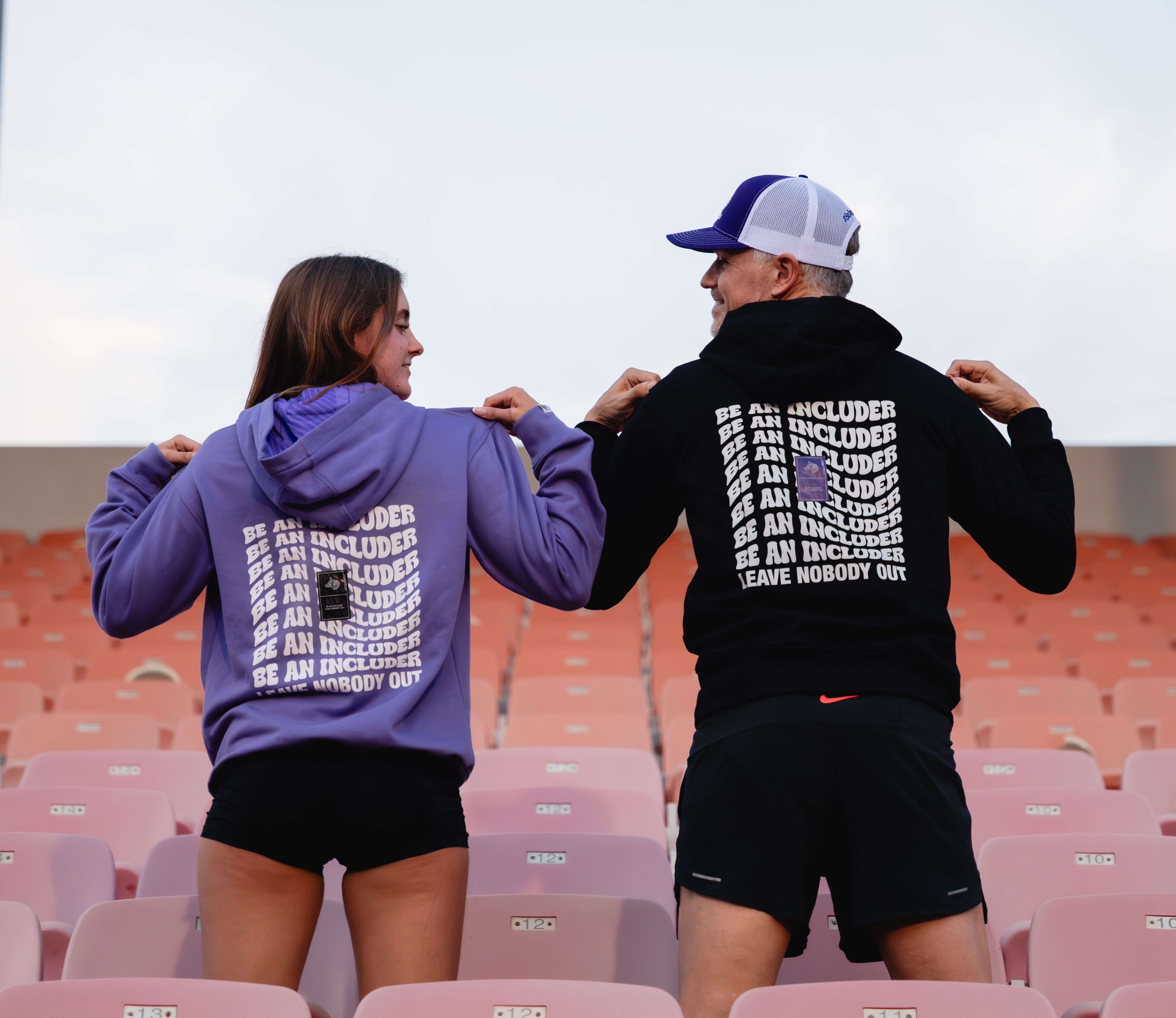 Two people wearing hoodies with "Be An Includer" messaging in an outdoor setting.