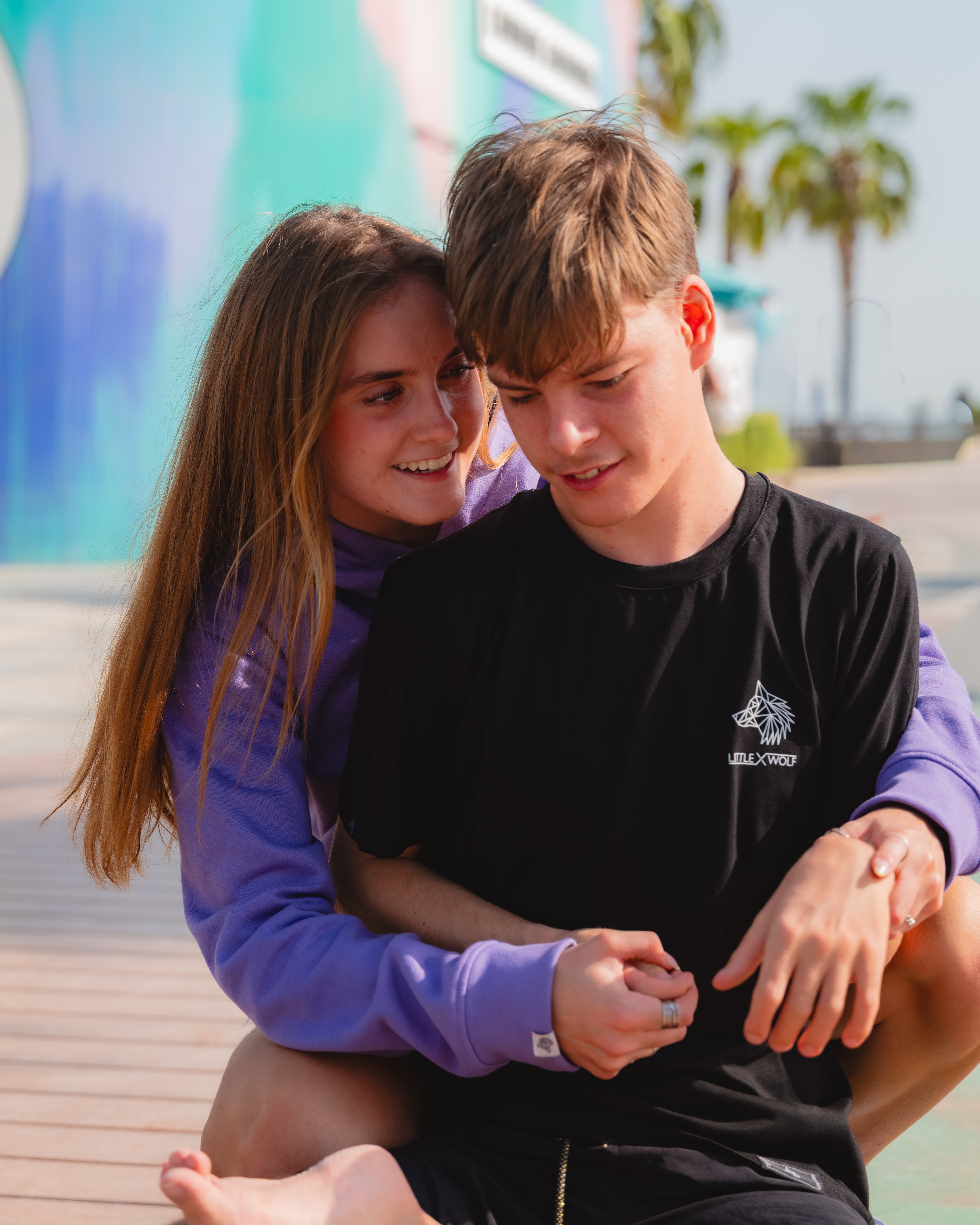 Rio & Tia sitting together on a skateboard with a colorful mural and palm trees in the background.