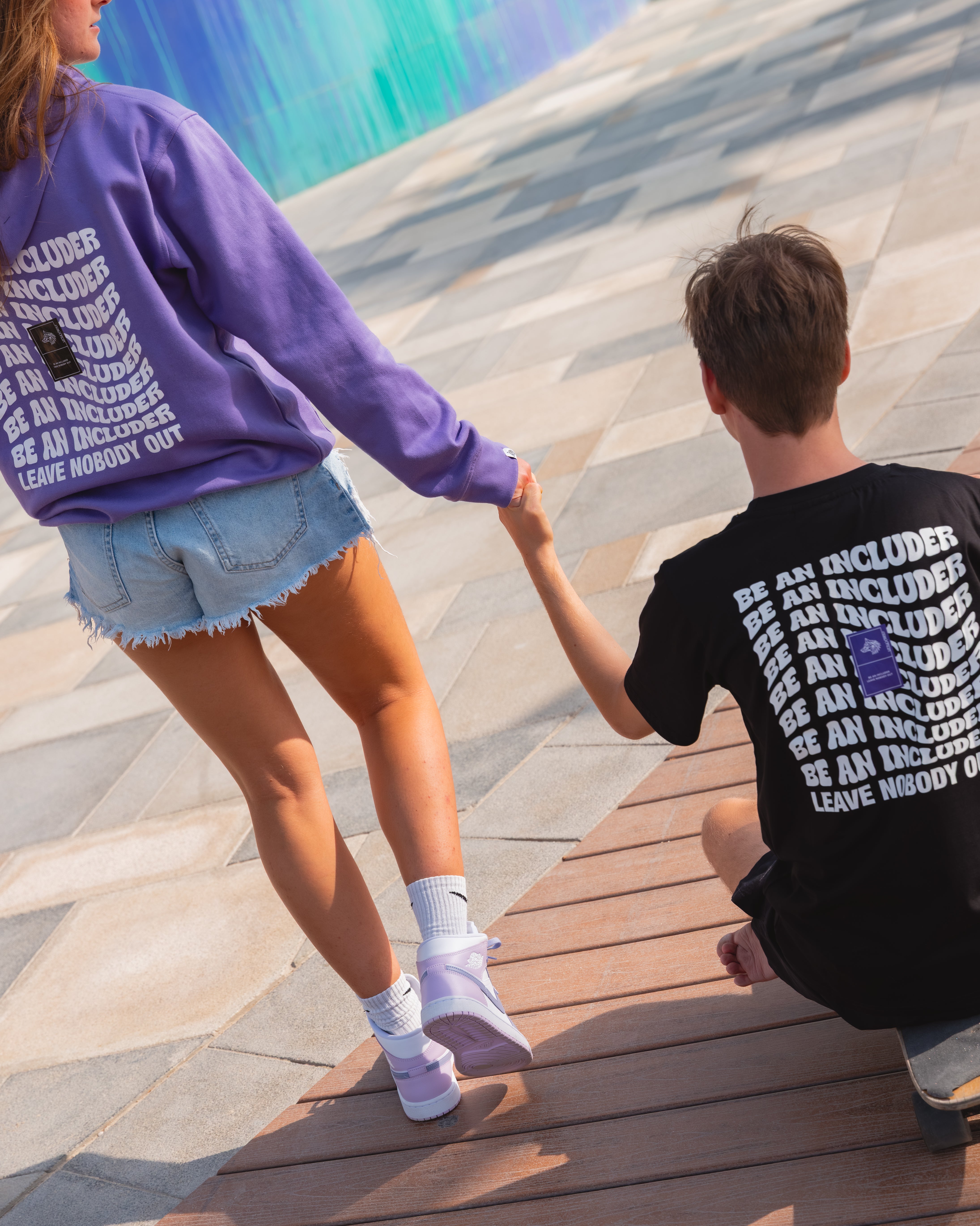 Rio & Tia holding hands on a skateboard with a colorful mural in the background.