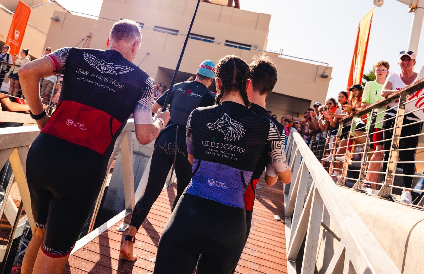 Athletes in wetsuits preparing to enter a pool, with spectators watching from the deck.